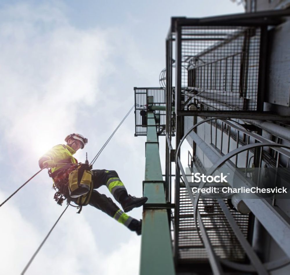 View from bottom on rope access technicians abseil from tower high up with sun behind